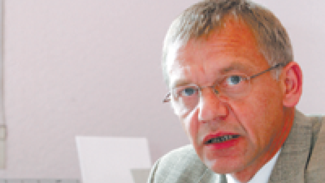 Vīrietis gaišā uzvalkā sēz pie rakstāmgalda