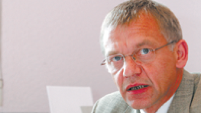Vīrietis gaišā uzvalkā ar brillēm sēž pie galda